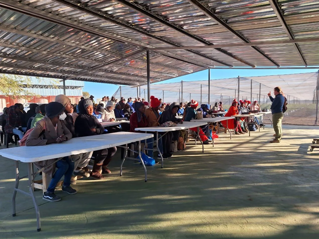 Gemeindemitglieder an langen Tischen unter einem Schattendach bei einem Treffen auf der Farm